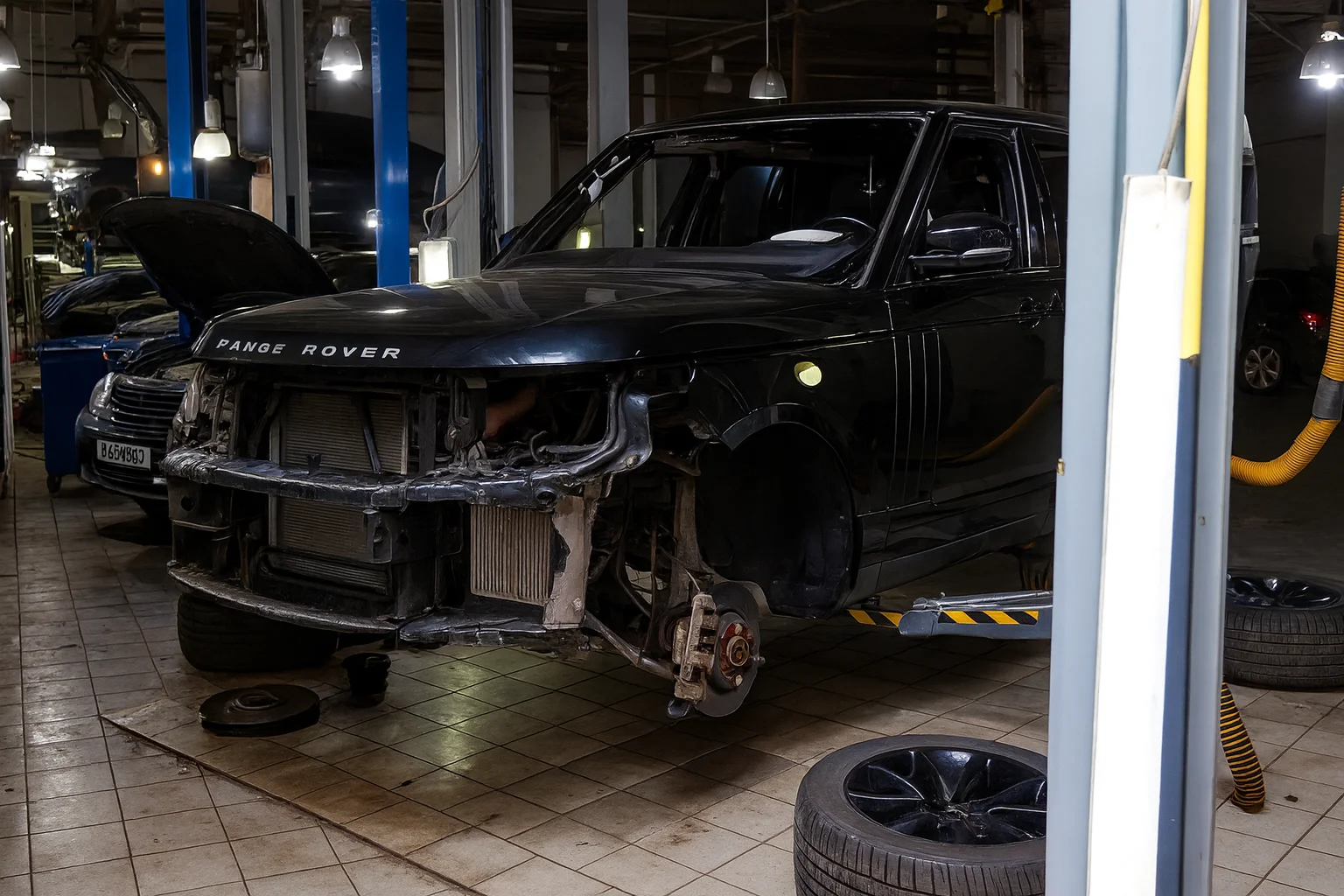 Black SUV inside an auto repair workshop with the front bumper removed, engine components exposed, and wheels taken off, surrounded by tools and tires on the garage floor under industrial lighting.