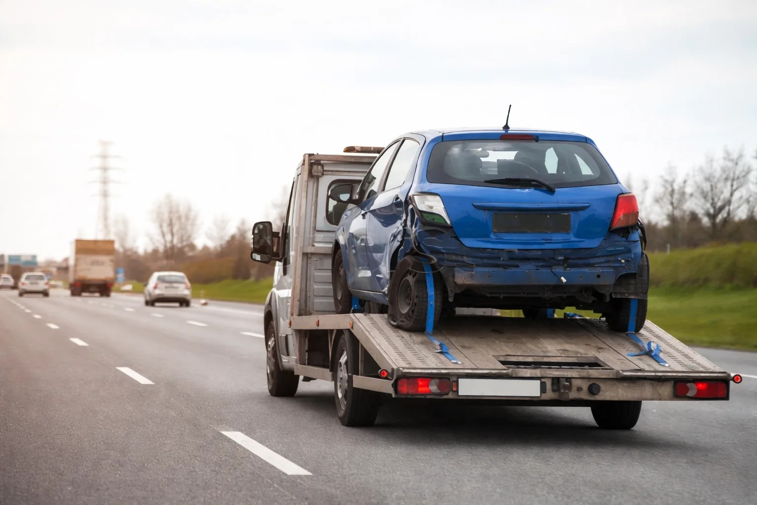 Tow truck transporting a damaged blue car on a highway, showcasing the best car recovery service providing safe and reliable vehicle towing after an accident.