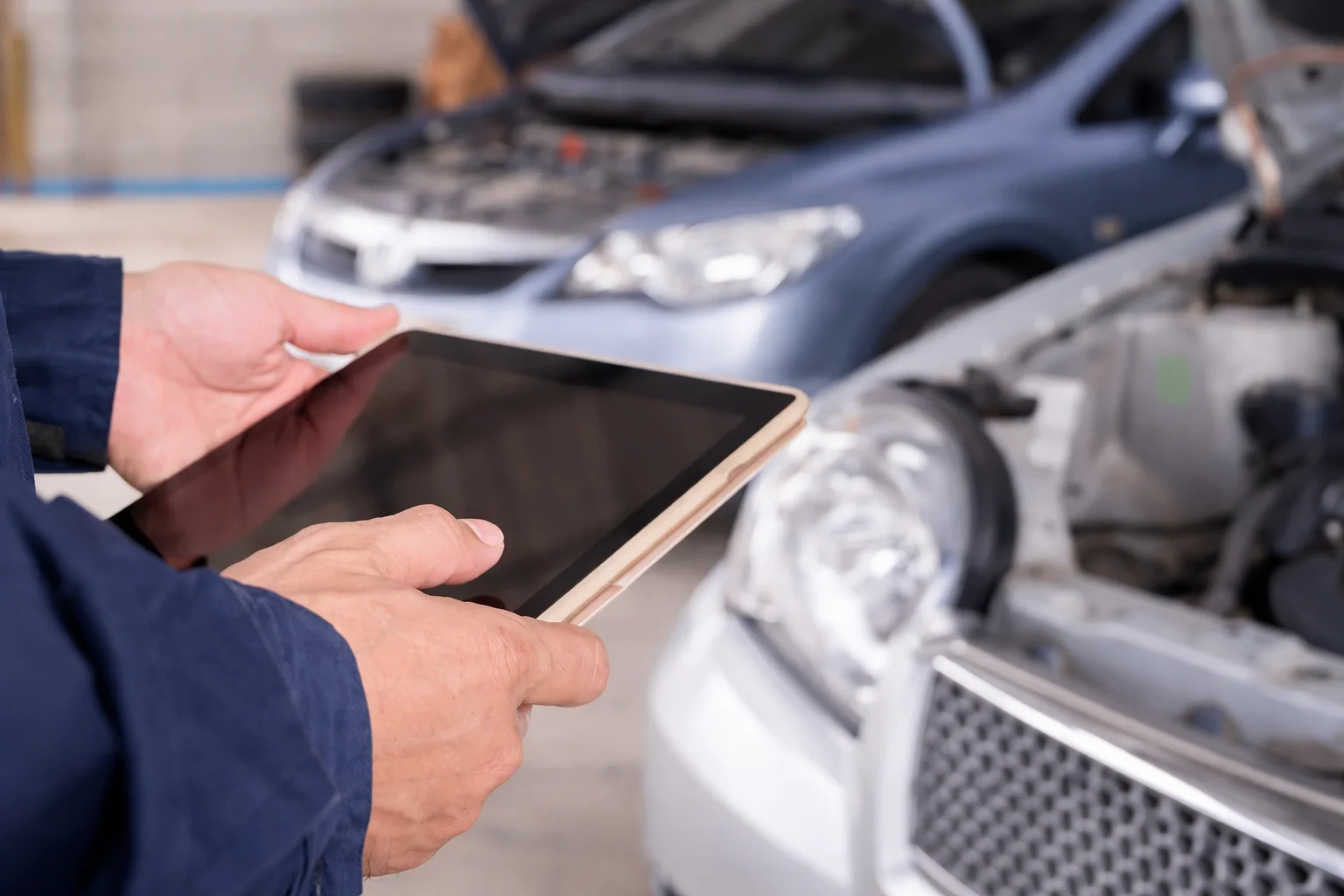 Technician holding a tablet while inspecting vehicles with open hoods inside a workshop, representing a professional Vehicle Inspection Center Near Me offering detailed digital diagnostics.