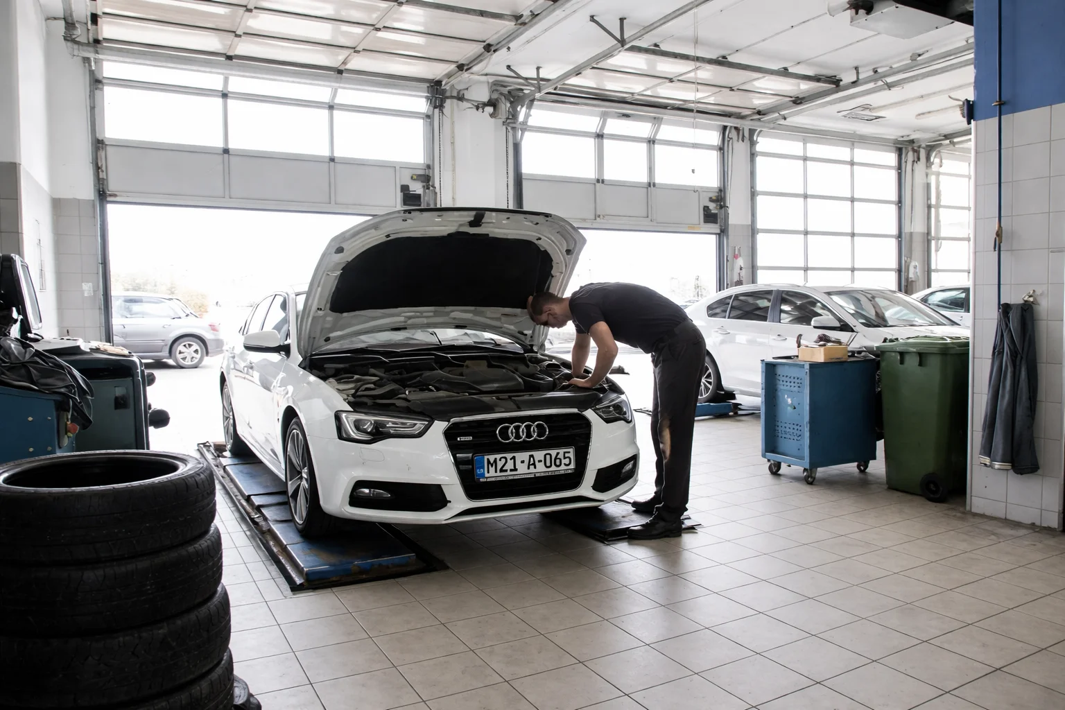 Technician inspecting a white Audi with the hood open inside a spacious service garage, illustrating professional maintenance at the nearest car workshop for reliable vehicle care.