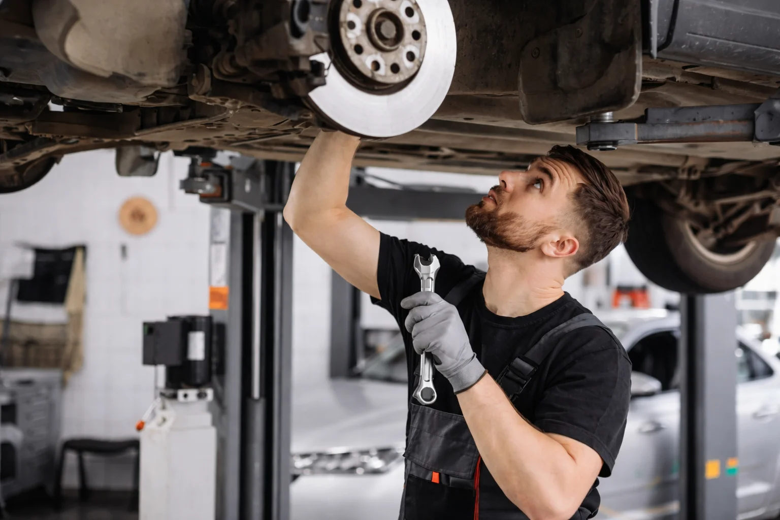 Professional auto mechanic inspecting and servicing the brake rotor and suspension under a lifted vehicle, performing detailed Mercedes brake repair in a modern automotive workshop.