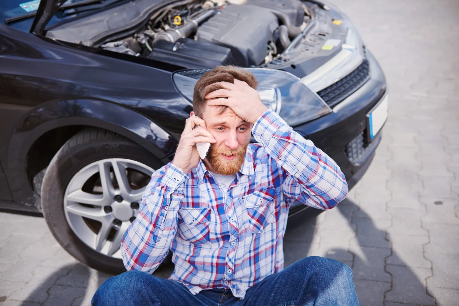Frustrated man sitting beside his car with the hood open, calling for roadside assistance after his car not starting unexpectedly on the roadside.
