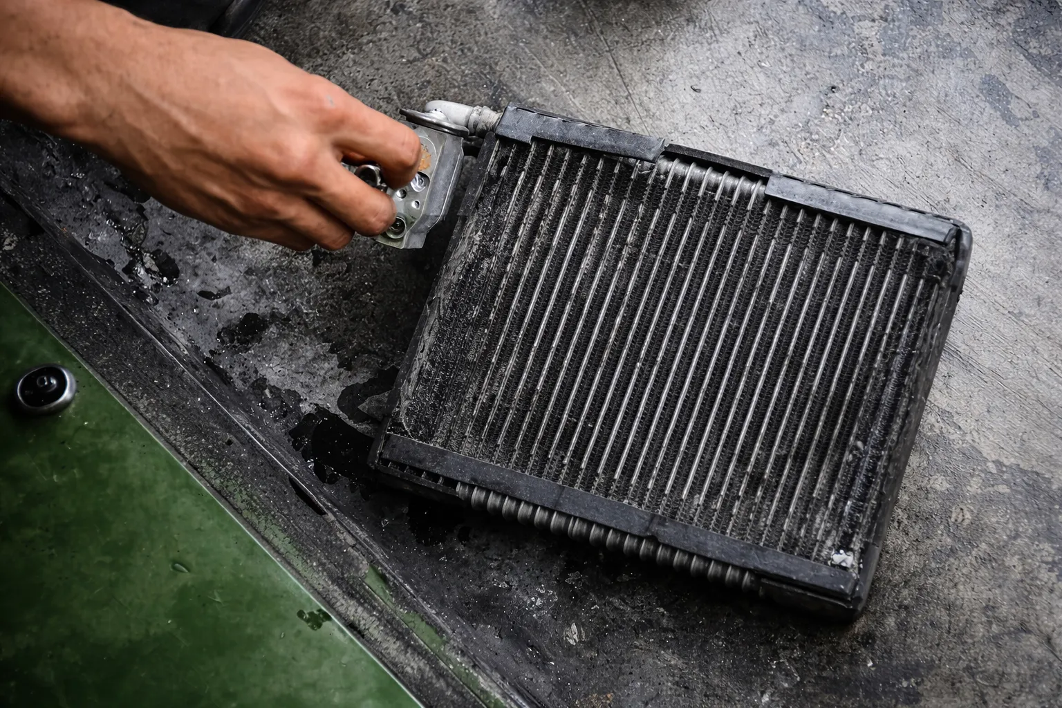 Close-up of a worn AC condenser on a workshop floor as a mechanic’s hand inspects the unit, illustrating a lamborghini AC condenser replacement to fix cooling issues and restore efficient air conditioning.