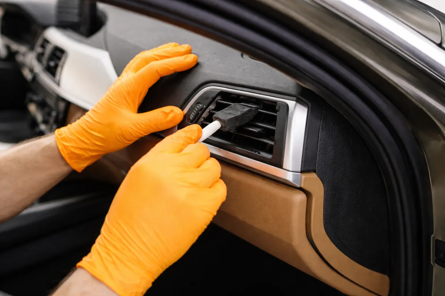 Close-up of a technician in orange gloves cleaning a car’s interior air vent with a detailing tool, illustrating careful lamborghini ac system flushing and ventilation cleaning for improved airflow and cabin comfort.