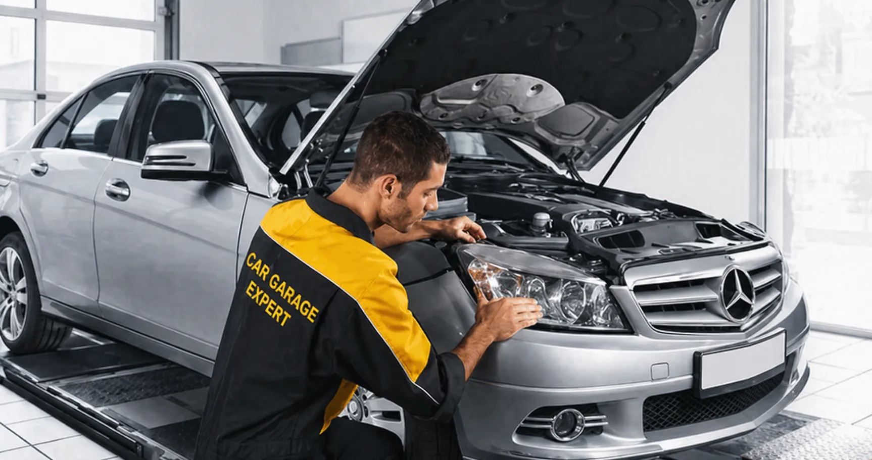 Technician in a black and yellow uniform labeled “CAR GARAGE EXPERT” inspecting the headlight of a silver Mercedes-Benz sedan with the hood open inside a modern workshop, showcasing a professional benz service center for expert vehicle maintenance and repair.
