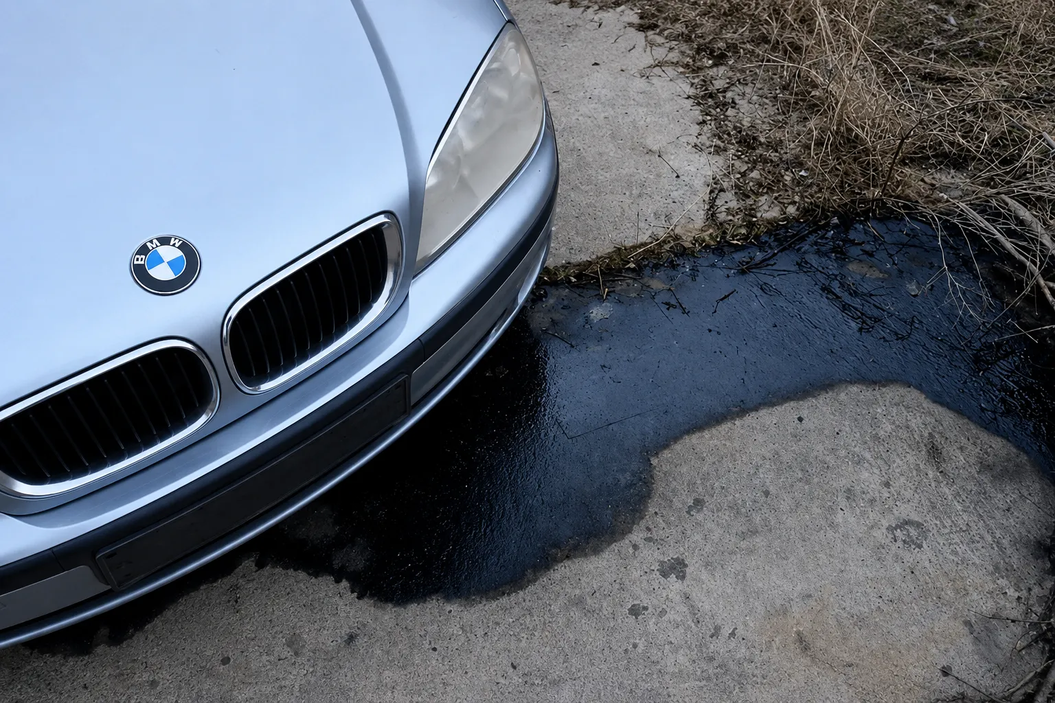 Close-up of a BMW vehicle with dark engine oil leaking onto the pavement, highlighting a common issue that requires professional BMW oil leaks repair to prevent engine damage and maintain vehicle performance.